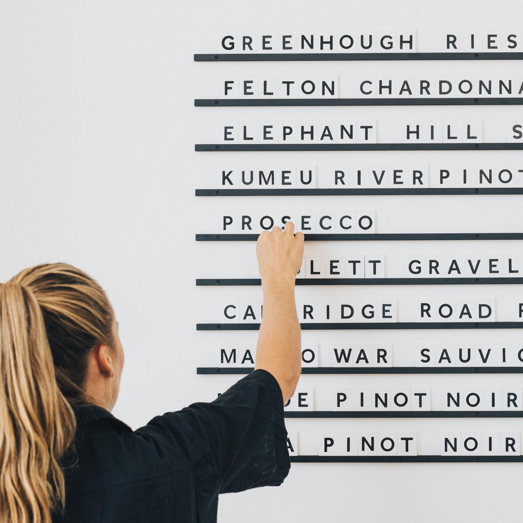 A girl adding wooden letter tiles to wall-mounted rails to display a wine menu on a white wall